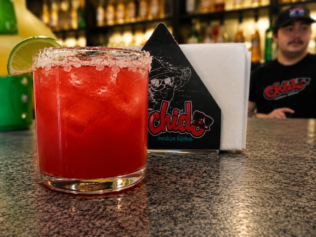 Vibrant fresh red house margarita cocktail in a salted-rim glass with lime wedge, next to Chido branded napkin holder