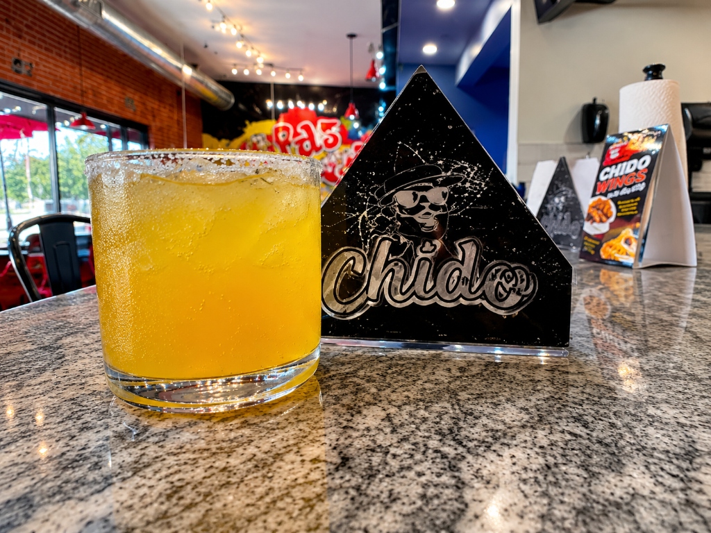 Vibrant fresh mango margarita cocktail in a salted-rim glass on counter, next to Chido branded sign